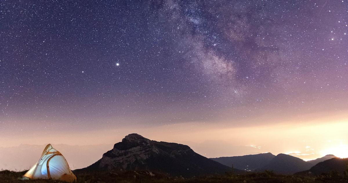 The Milky Way galaxy lights up the night sky as a tent with light on is visible in the foreground.