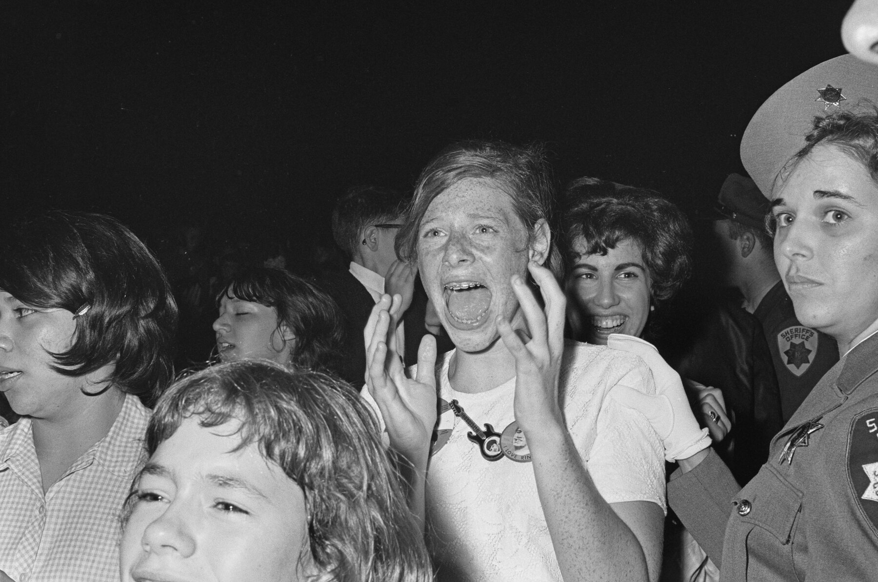 A black-and-white photo of Beatles fans screaming in a  crowd, with a security officer placing a white-gloved hand on the shoulder of one screeching girl.
