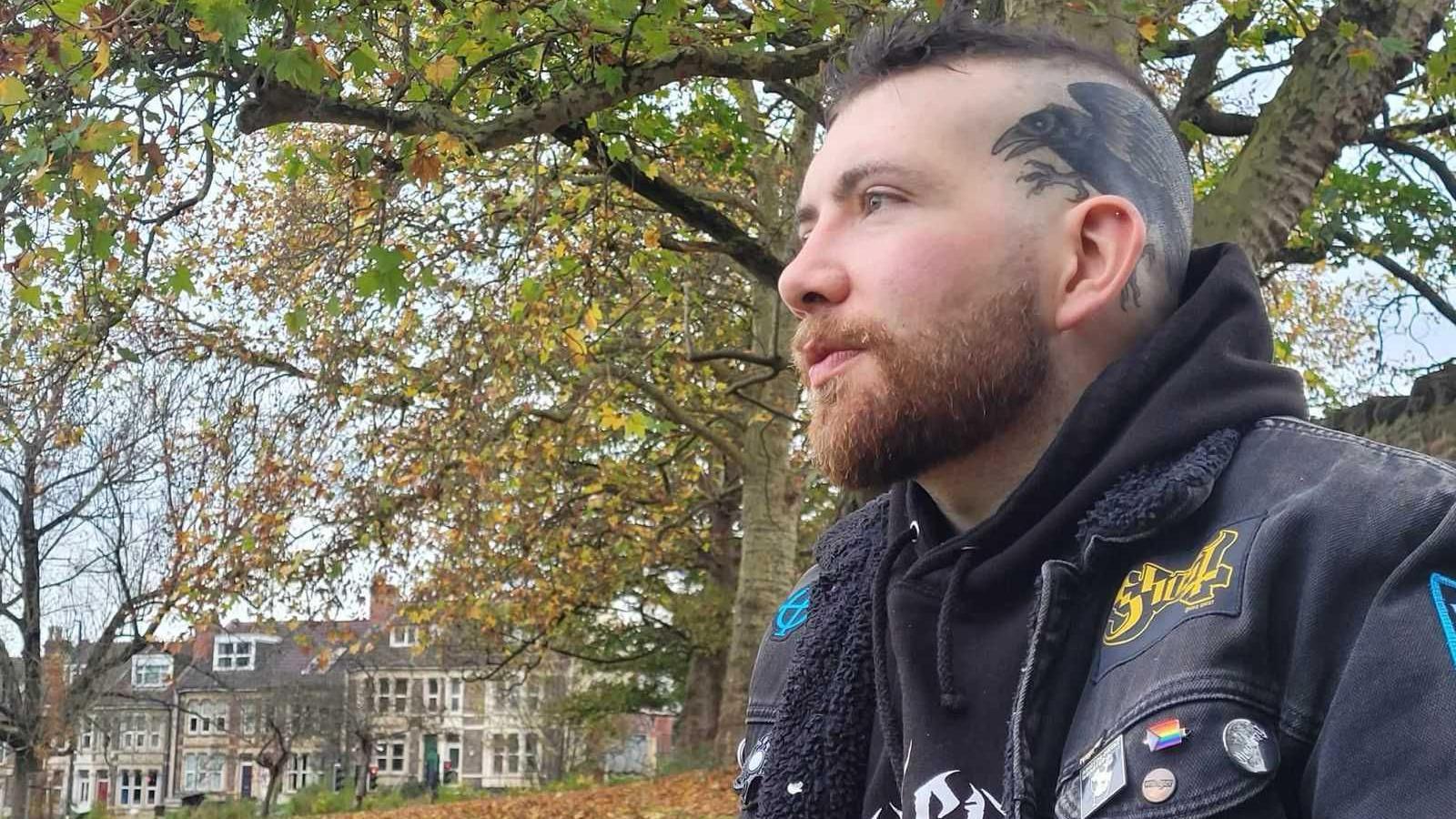 Patrick Darling wearing a black hoodie underneath a black denim jacket, decorated with various colourful pins and patches. He has dark hair which is shaved on the sides, and a tattoo of a raven across the left side of his head. He is sitting outdoors in a park surrounded by trees, looking off to the left of the camera. In the background there is a row of terraced houses.