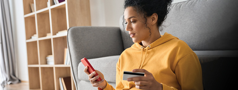A woman in a yellow hoodie sits on the floor in a living room setting, holding a credit card and a smartphone, likely making an online purchase. There is a wooden bookshelf and gray couch in the background.
